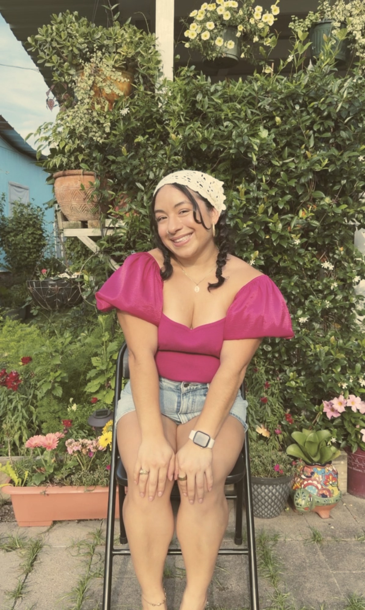 Eveling sitting in a lush garden surrounded by plants and flowers, smiling, wearing a magenta top and a cream bandana