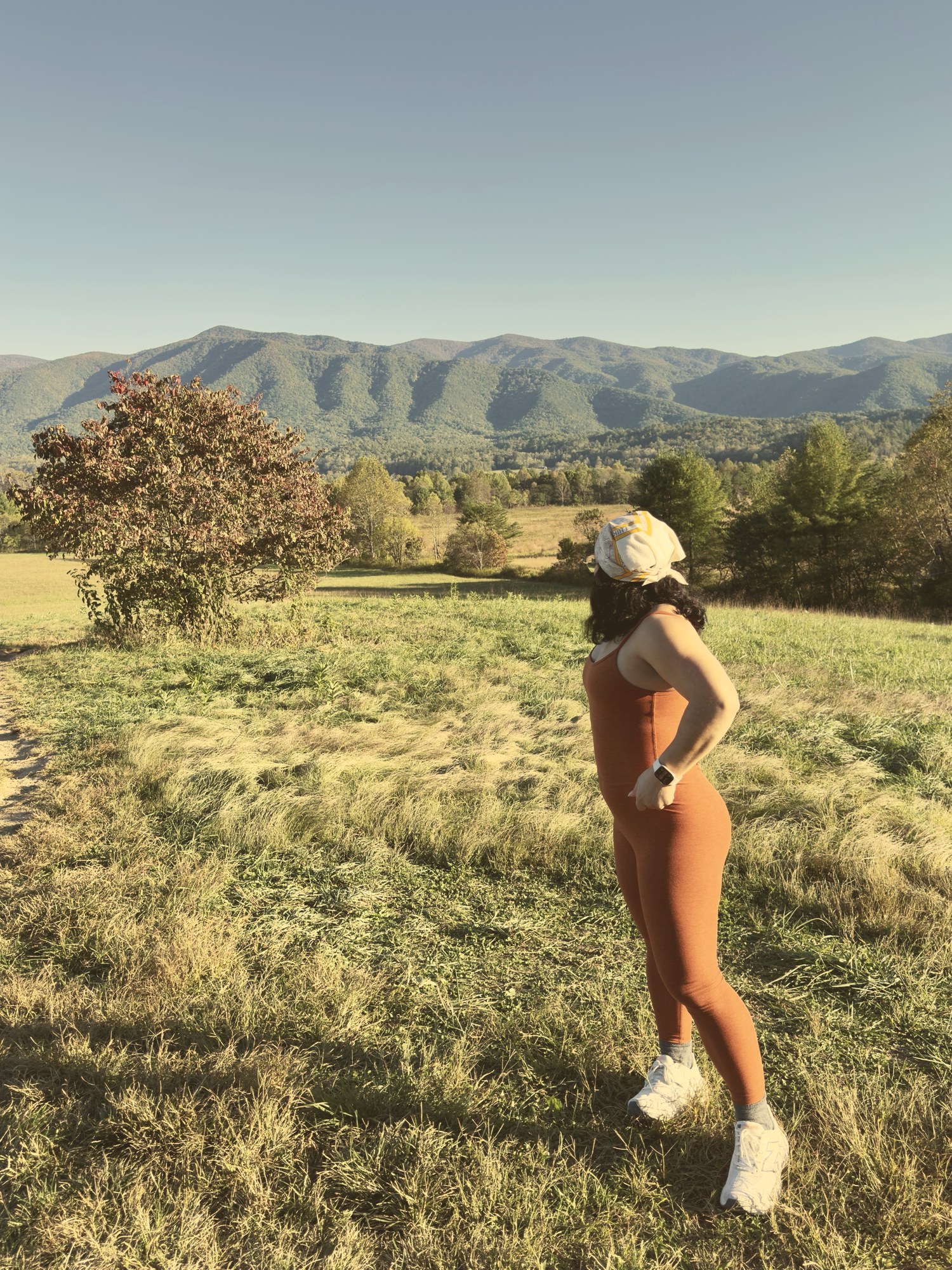 Eveling standing in a golden mountain meadow in a terracotta outfit, looking out at the mountains